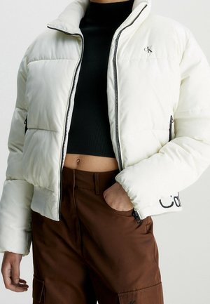 Personne portant une veste matelassée blanche avec une fermeture éclair noire sur un haut court noir et un pantalon marron, main dans la poche.