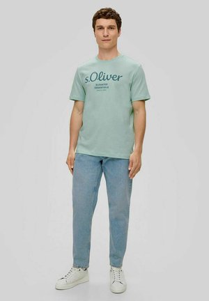 Mintgrünes T-Shirt mit Rundhalsausschnitt und Frontdruck, kombiniert mit hellblauen Jeans und weißen Sneakers. Aus glattem Baumwollstoff.