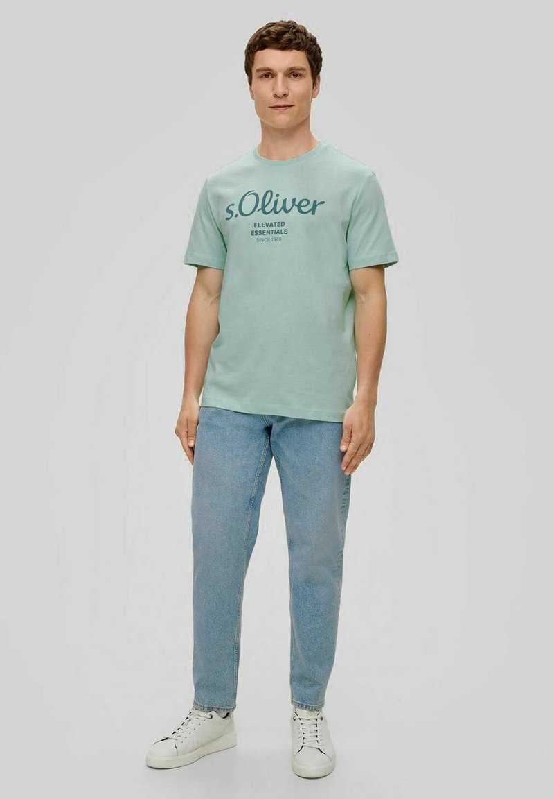 Mintgrünes T-Shirt mit Rundhalsausschnitt und Frontdruck, kombiniert mit hellblauen Jeans und weißen Sneakers. Aus glattem Baumwollstoff.