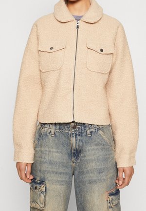 Beige Fleecjacke mit rundem Kragen, vorderen Taschen und Reißverschluss. Trägt sie über Jeans im Denim-Stil mit einem ausgewaschenen, abgenutzten Muster.