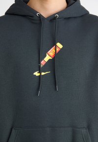 Mørkegrøn hættetrøje med et grafisk motiv af et gult rør med en rød label og et gult swoosh. Lavet af tekstureret stof med en frontlomme.