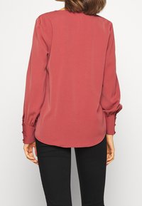 Langärmlige, burgunderfarbene Bluse mit glatter Textur und gerafften Manschetten, die mit kleinen Knöpfen versehen sind. Runder Ausschnitt mit einem lässigen Schnitt.