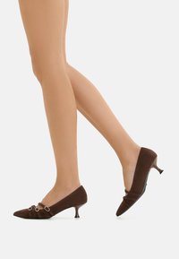 Scarpe con tacco alto in suede marrone con punte affusolate, dotate di anelli decorativi e un piccolo tacco kitten. Texture liscia e design minimalista.