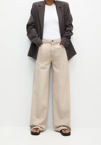 Jean large beige taille haute, associé à un haut blanc ajusté et un blazer oversize gris foncé. Texture du tissu lisse.