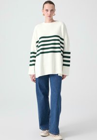 Pulover oversized alb din tricot cu dungi orizontale verzi închise, asortat cu blugi Levis albaștri cu picior larg și pantofi bej.