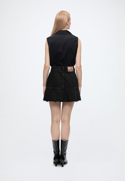 Femme aux longs cheveux blonds portant un haut noir sans manches, une jupe plissée noire avec un patch, et des bottes noires à talons, debout de dos.