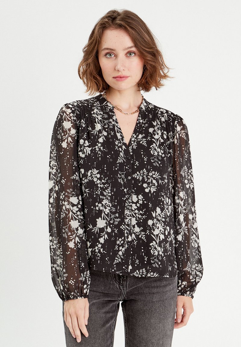 Blouse noire avec un motif floral blanc, manches longues transparentes, décolleté en V, boutons à l'avant et poignets élastiques. Associée à un jean gris foncé.