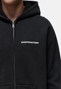 Felpa nera con zip realizzata in materiale morbido, caratterizzata da un logo frontale "GOODFORNOTHING" in bianco. Il design include un cappuccio con cordoncino e tasche frontali.