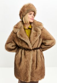 Brun faux fur-frakke med bred krave, bælte i taljen og matchende pelsvinterhue. Har en blød tekstur og et tofarvet designmønster.