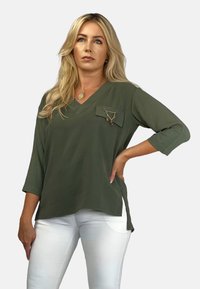 Oliwkowy, top w dekolcie V wykonany z lekkiego materiału, z kieszenią na piersi z łańcuszkowymi akcentami i rękawami do 3/4. Połączony z białymi spodniami.