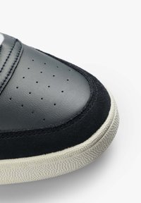 Schwarze Ledersneaker mit Wildlederakzenten, perforiertem Zehenbereich, weißen Nähten und einer strukturierten, cremefarbenen Sohle. Eingearbeitete geschwungene Designelemente.