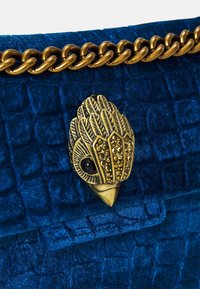 Modrá sametová kabelka s texturovaným povrchem, vybavená zlatým řetízkovým popruhem a podrobným uzávěrem ve tvaru ptáka zdobeným černými a zlatými akcenty.