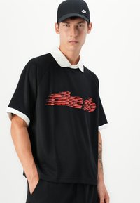 Black polo shirt with white collar and short sleeves, featuring a red striped logo across the chest. Smooth fabric with a relaxed fit.
