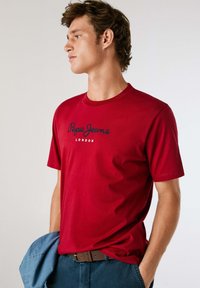 Raudona medvilninė marškinė su apvaliu kaklu ir trumpomis rankovėmis, su juodu užrašu "Pepe Jeans London" priekyje.