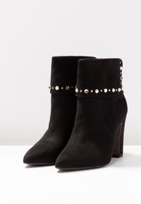 Stivali alla caviglia in suede nero con punta affusolata e tacco alto a cono. Presentano una fascia decorativa di borchie attorno al gambale superiore.