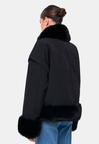 Giacca nera con colletto e polsini in pelliccia sintetica. Presenta un design squadrato e una texture liscia e opaca. Cuciture visibili lungo il dietro.