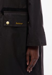 Fekete hosszú kabát oldalzsebbel, sárgaréz patentgombbal, bordás részletekkel és arany színű "Barbour" hímzéssel. Simára szőtt anyagtextúra.