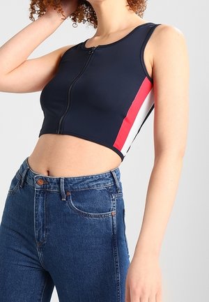 Femme portant un débardeur court bleu marine avec une fermeture éclair devant et des bandes rouges et blanches sur les côtés, associé à un jean bleu taille haute.