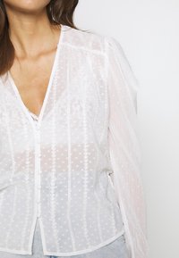 Chemisier blanc transparent avec pois texturés et broderie rayée délicate ; doté d'un décolleté en V et d'une fermeture boutonnée à l'avant.