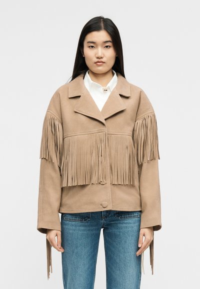 Vrouw draagt een beige suède jas met franjes over de borst en armen, een wit shirt eronder en een blauwe spijkerbroek, staande tegen een witte achtergrond.