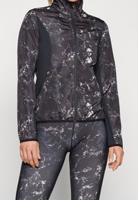 Černá sportovní bunda s mramorovým vzorem, vysokým límcem, plným zipem a kontrastními černými bočními panely; přiléhavý střih a elastické manžety.