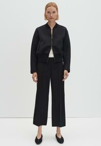 Blouson bombardier noir à ourlet raccourci, fermeture éclair devant et poignets côtelés, associé à un pantalon noir à jambes larges et des chaussures plates. Design minimaliste.