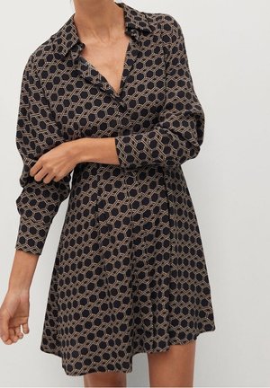 Kvinde iført en langærmet, sort kjole med et beige geometrisk sekskantmønster, krave og taljeret pasform.
