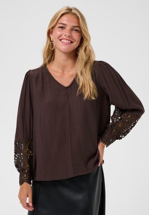 Lachende vrouw met lang blond haar, die een donkerbruine blouse met kanten mouwen en een zwarte rok draagt tegen een effen achtergrond.