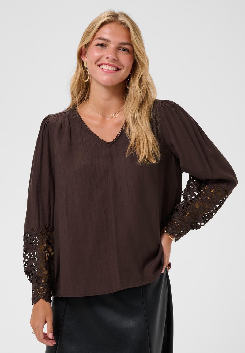 Femme souriante aux longs cheveux blonds, portant un chemisier marron foncé avec des manches en dentelle et une jupe noire sur un fond uni.