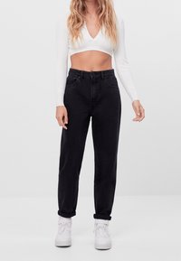 Jeans negros de talle alto con un corte holgado, que cuentan con cinco bolsillos y puños doblados, combinados con una blusa corta de manga larga blanca y zapatillas blancas.