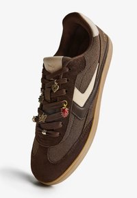 Brązowe zamszowe sneakersy z beżowymi akcentami, z ozdobnymi charmsami na sznurowadłach i gumową podeszwą. Okrągły czubek i wyściełany kołnierz.
