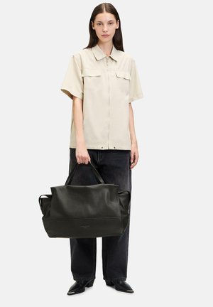 Schwarze Leder-Shopper-Tasche mit strukturierter Oberfläche, geräumigem Design und robusten Griffen, zusammen mit einem Modell, das ein beiges Hemd und dunkle Jeans trägt.