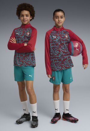 Twee jonge voetballers in Portugal-tenues staan naast elkaar; een van hen houdt een voetbal onder zijn arm, beiden dragen voetbalschoenen en witte sokken.