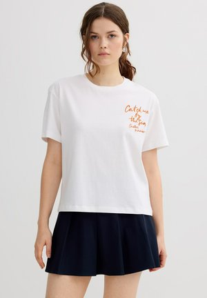 Joven con cabello castaño que lleva una camiseta blanca con texto naranja en el lado izquierdo del pecho y una falda plisada negra, de pie contra un fondo liso.