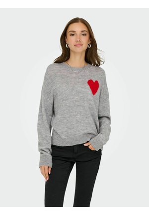 Grå stickad tröja med lång ärm, med ett texturerat röd hjärtdesign på vänster bröst. Bärs med svarta byxor.