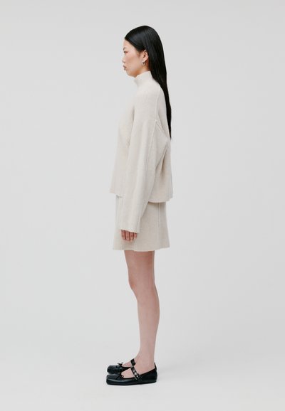 Maglione a coste beige con collo alto, abbinato a una gonna a coste coordinata. Scarpe basse nere con dettagli a strappo. Vista laterale.