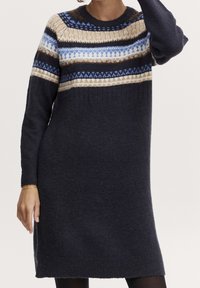 Femme portant une robe-pull en tricot à manches longues avec des rayures à motifs bleu marine, beige, marron et bleu clair traversant la poitrine et les manches.