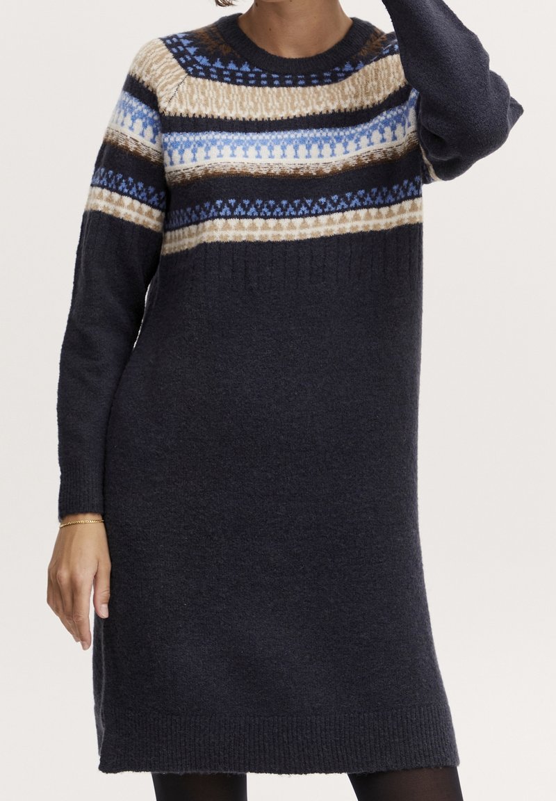 Femme portant une robe-pull en tricot à manches longues avec des rayures à motifs bleu marine, beige, marron et bleu clair traversant la poitrine et les manches.
