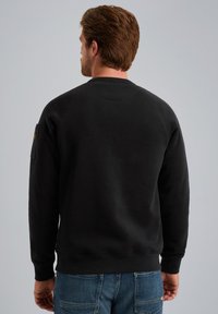 Sudadera negra fabricada con una mezcla de algodón, presenta un cuello redondo y puños acanalados. Incluye un bolsillo con parche en el brazo izquierdo. Llevada con jeans azules.