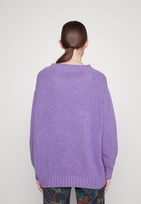Personne avec une queue de cheval brune portant un pull violet surdimensionné et texturé ainsi qu'un pantalon à motifs floraux, vue de dos sur un fond clair uni.