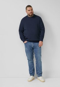 Granatowa teksturowana bluza z okrągłym dekoltem, zestawiona z jasnoniebieskimi dżinsami i białymi trampkami. Model stoi na jednolitym tle.