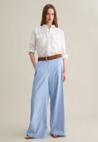 Camisa de lino blanca con dos bolsillos en el pecho, combinada con pantalones de pierna ancha a rayas finas en azul claro y un cinturón marrón con una hebilla circular.