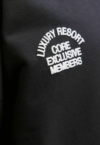 Tejido negro con texto bordado en blanco: "RESORT DE LUJO", "NÚCLEO" y "MIEMBROS EXCLUSIVOS" en formación de arco. Textura suave.