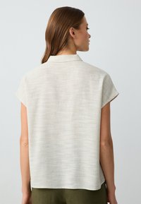 Blusa corta a maniche corte beige chiaro con un tessuto leggermente testurizzato, dotata di colletto e una vestibilità ampia che arriva all'orlo.