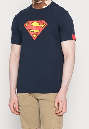 Uomo con maglietta blu navy con grande logo di Superman rosso e giallo e pantaloncini cargo beige, in piedi davanti a uno sfondo semplice.