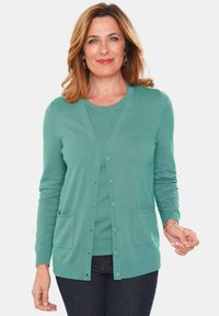 Strickjacke in Türkis mit V-Ausschnitt, Knopfverschluss und zwei vorderen Taschen. Lange Ärmel mit gerippten Bündchen und Saum. Kombiniert mit einem passenden Oberteil.