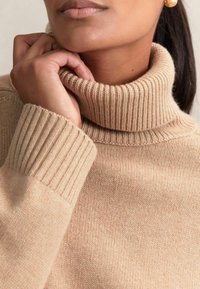 Beige coltrui gemaakt van zacht gebreid materiaal, met geribbelde manchetten en kraag, met een gladde textuur en een relaxed fit.