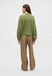 Πράσινο πλεκτό πουλόβερ με χαμηλωμένους ώμους, ριμπ τελείωμα και μπεζ παντελόνι με ευρεία μπατζάκια, κατασκευασμένο από ύφασμα με ομαλή υφή.