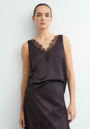 Donna che indossa un top nero in raso senza maniche con bordatura in pizzo e gonna coordinata con dettagli in pizzo, in piedi contro uno sfondo bianco semplice.