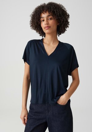Jeune femme aux cheveux bouclés portant un haut bleu foncé à manches courtes avec un col en V et un jean foncé, debout avec une main dans la poche.